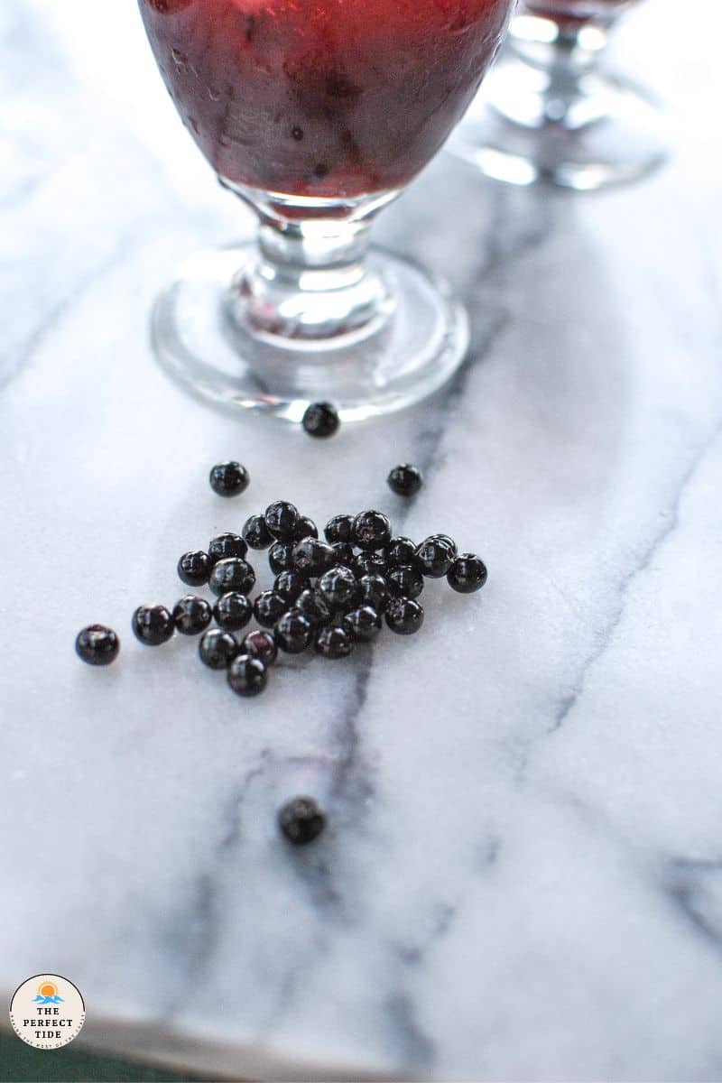 fresh huckleberries on marble countertop next to huckleberry margarita in clear gobler glass