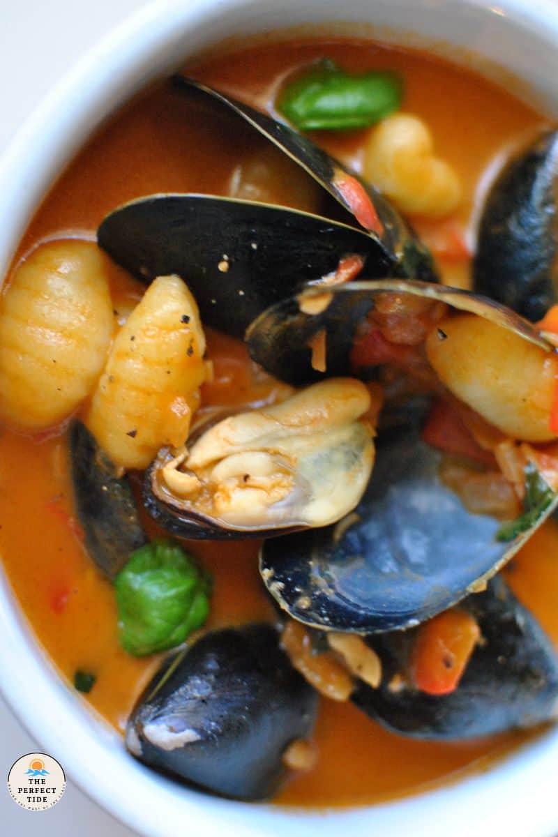 Mussel soup in tomato sauce with roasted red pepper, gnocchi, and basil in white bowl