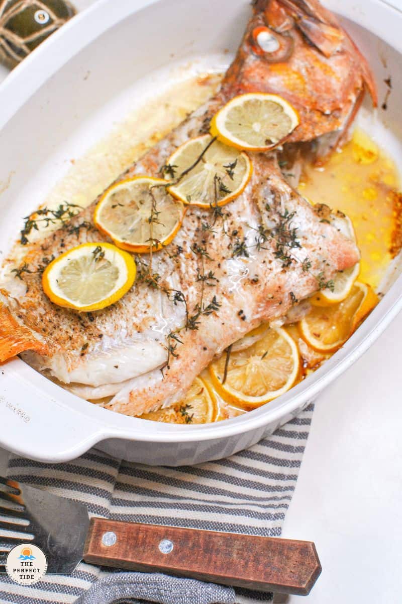 baked whole rockfish with lemon and thyme in white baking dish fish spatula and striped black and white napkin