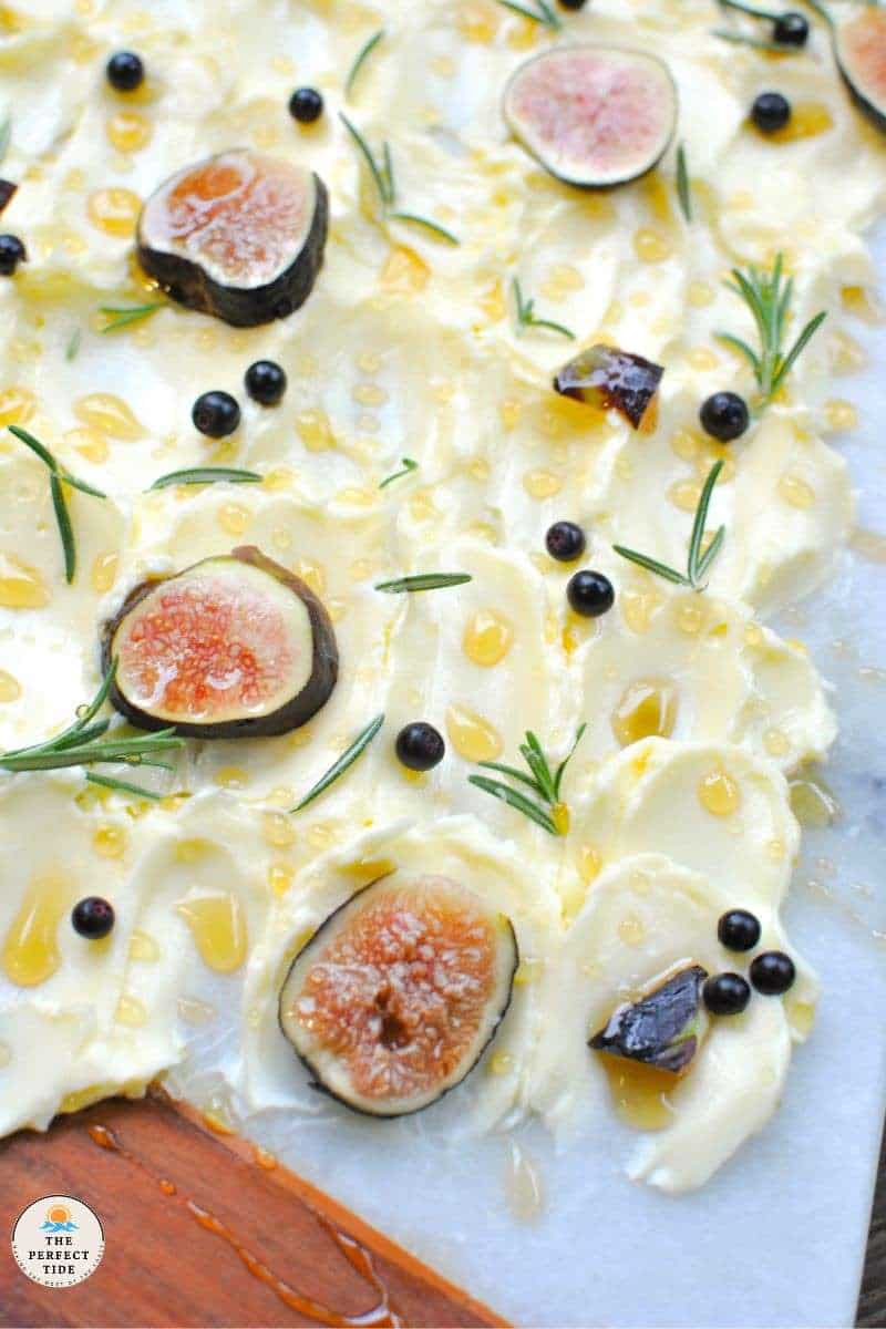 close up of fig and rosemary on a butter board with butter smeared on a marble and wood board with rosemary fresh figs sliced, honey fresh huckleberries and spicy honey 