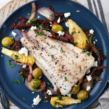 Black cod baked with olives artichokes red onion tomatoes feta on blue plate