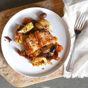 Mediterranean baked fish with artichokes olives sun-dried tomatoes on white plate