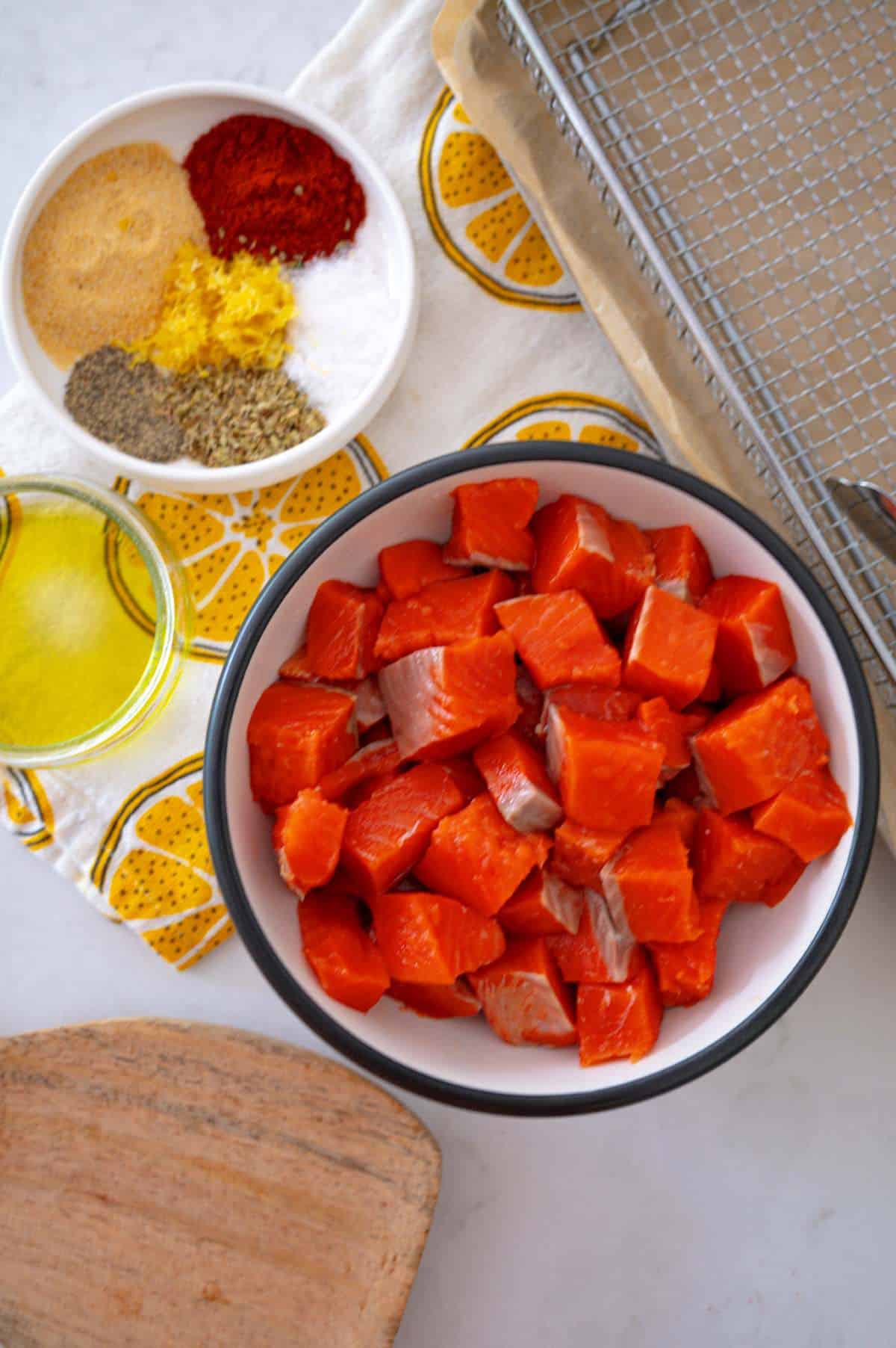 ingredients to make air fryer salmon bites. bowl of salmon pieces near a plate of spices and olive oil. lemon towel nearby